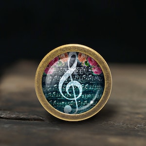 May include: A round, brass-coloured pendant with a glass insert featuring a white treble clef on a teal background with musical notes and pink flowers.