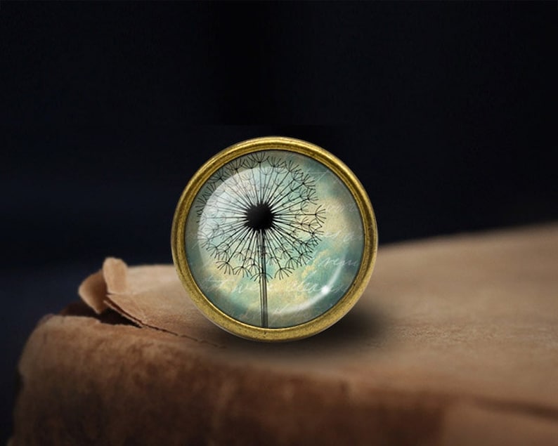 Op de afbeelding: Een ronde, goudkleurige hanger met een glazen cabochon met een zwart paardenbloem silhouet tegen een blauwe en witte aquarel achtergrond.