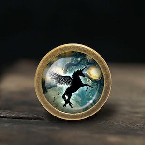 May include: A round, brass-colored pendant with a glass dome featuring a black silhouette of a winged unicorn against a blue and green forest background.