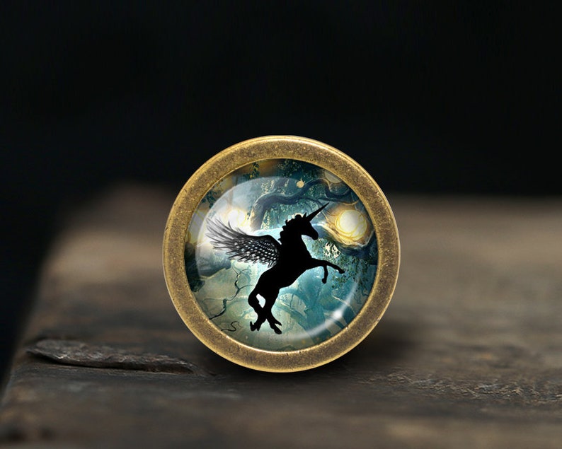 Pode incluir: Um pingente redondo de cor lat&atilde;o com uma c&uacute;pula de vidro que apresenta uma silhueta preta de um unic&oacute;rnio alado sobre um fundo de floresta azul e verde.