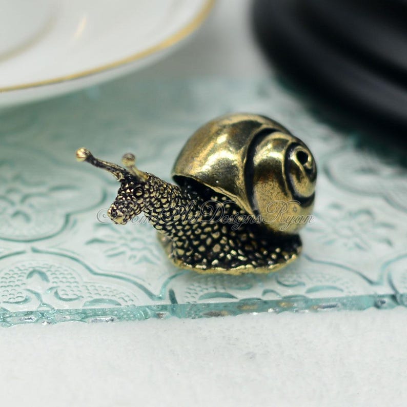 Puede incluir: Una peque&ntilde;a figurita de caracol de color dorado con una concha y un cuerpo detallados. El caracol est&aacute; sentado sobre una superficie de vidrio transparente con un patr&oacute;n decorativo.