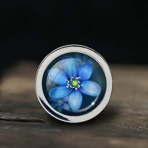 Puede incluir: Un anillo de plata con una flor azul encerrada en vidrio. La flor tiene cinco p&eacute;talos y un centro amarillo.