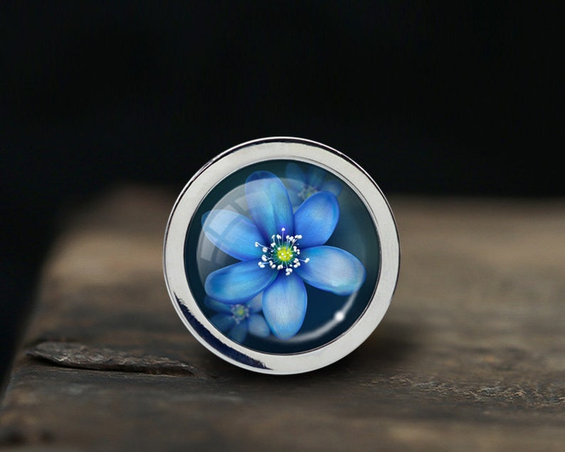 Puede incluir: Un anillo de plata con una flor azul encerrada en vidrio. La flor tiene cinco p&eacute;talos y un centro amarillo.