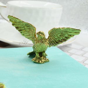 Pode incluir: Uma figura de águia dourada com as asas abertas. A águia está pousada sobre uma superfície turquesa.