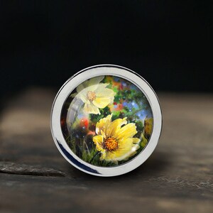 May include: A silver ring with a glass dome featuring a painted image of yellow flowers with green leaves and a blue background.