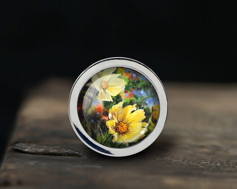 May include: A silver ring with a glass dome featuring a painted image of yellow flowers with green leaves and a blue background.