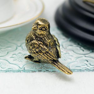 May include: A detailed, antique-style brass bird figurine. The bird is perched, with textured feathers and a detailed beak. The figurine is a warm gold color, and sits on a clear, patterned glass surface. The background includes a white plate with a gold rim.