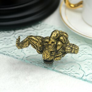 Puede incluir: Un pomo de armario de metal dorado con forma de mono sentado con las piernas cruzadas. El mono est&aacute; detallado con pelo y tiene una expresi&oacute;n realista.