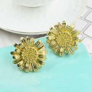 May include: A pair of gold sunflower earrings. The earrings are made of metal and feature a detailed sunflower design.