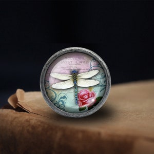 K&ouml;nnte beinhalten: Ein runder silberfarbener Knopf mit einem Glaseinsatz, der ein Vintage-Postkarten-Design zeigt. Die Postkarte hat einen rosafarbenen Hintergrund mit einer wei&szlig;en Libelle und einer rosafarbenen Rose. Der Text auf der Postkarte lautet "Souvenir Post Card".