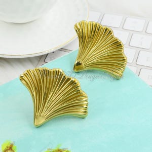 May include: A pair of gold-tone earrings shaped like ginkgo leaves. The earrings are made of metal and have a shiny finish.