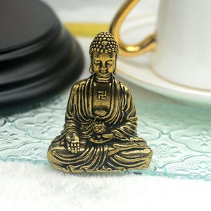 Peut inclure: Une petite statue de Bouddha dorée assise en position de méditation, les mains jointes devant la poitrine. La statue a un visage détaillé et des vêtements complexes.