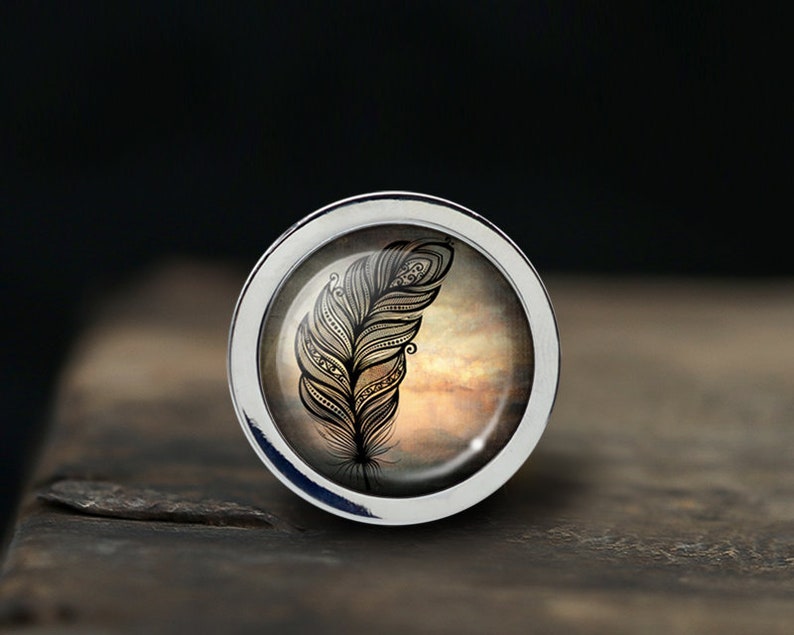 May include: A silver ring with a glass cabochon featuring a black and white detailed feather design on a brown and orange background.