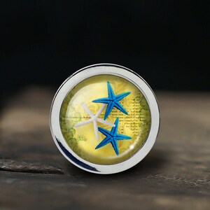 May include: A silver-toned round pendant with a glass dome containing three starfish. Two of the starfish are blue and one is white. The background is a yellow and green design with faded text.