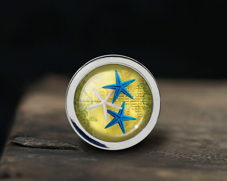 May include: A silver-toned round pendant with a glass dome containing three starfish. Two of the starfish are blue and one is white. The background is a yellow and green design with faded text.