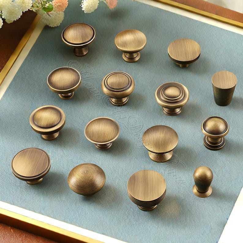 Puede incluir: Una colecci&oacute;n de tiradores de armario de lat&oacute;n antiguo en varias formas y tama&ntilde;os. Los tiradores tienen un acabado c&aacute;lido y envejecido, con dise&ntilde;os planos, redondeados y estriados. El hardware se muestra sobre una superficie azul claro, sugiriendo accesorios decorativos para el hogar.