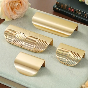 K&ouml;nnte beinhalten: Vier goldfarbene Schrankgriffe in verschiedenen Gr&ouml;&szlig;en. Zwei Griffe haben ein durchgehendes, gebogenes Design, w&auml;hrend die anderen beiden ein blattartiges Ausschnittmuster aufweisen. Die Griffe sind auf einer hellgr&uuml;nen Oberfl&auml;che angeordnet.