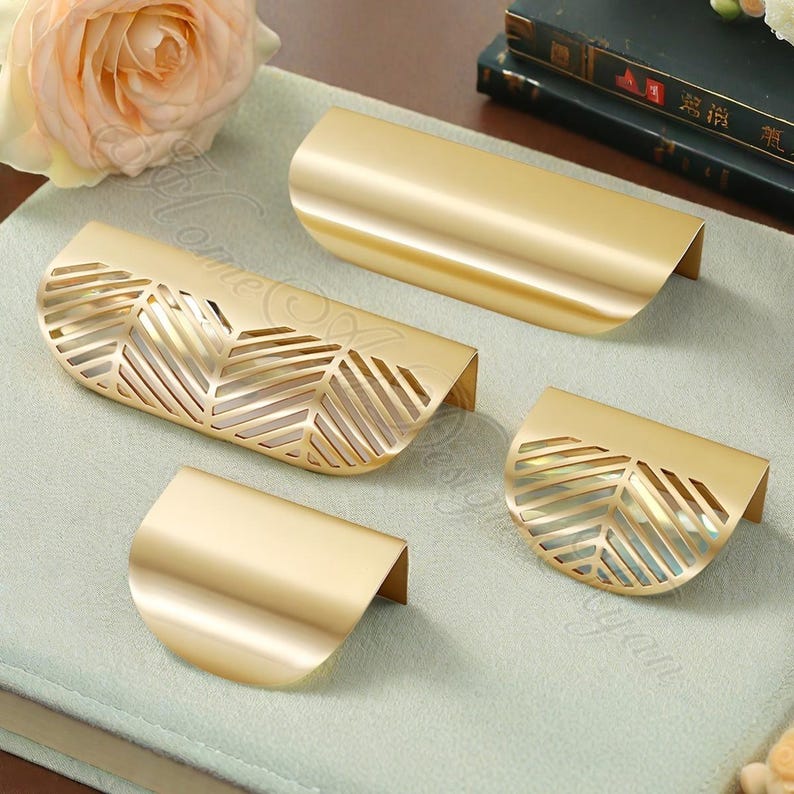 K&ouml;nnte beinhalten: Vier goldfarbene Schrankgriffe in verschiedenen Gr&ouml;&szlig;en. Zwei Griffe haben ein durchgehendes, gebogenes Design, w&auml;hrend die anderen beiden ein blattartiges Ausschnittmuster aufweisen. Die Griffe sind auf einer hellgr&uuml;nen Oberfl&auml;che angeordnet.