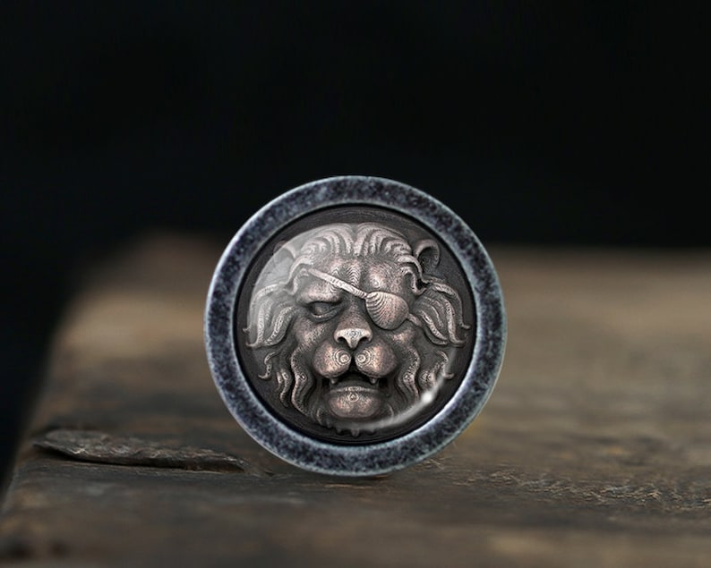 Puede incluir: Un bot&oacute;n redondo plateado con un dise&ntilde;o detallado de cabeza de le&oacute;n. El le&oacute;n tiene un parche negro sobre su ojo derecho.