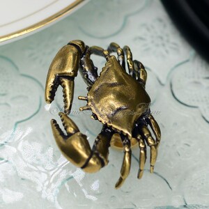 Puede incluir: Una figurita de cangrejo de metal dorado con garras y patas detalladas. El cangrejo est&aacute; colocado sobre una superficie texturizada azul claro.
