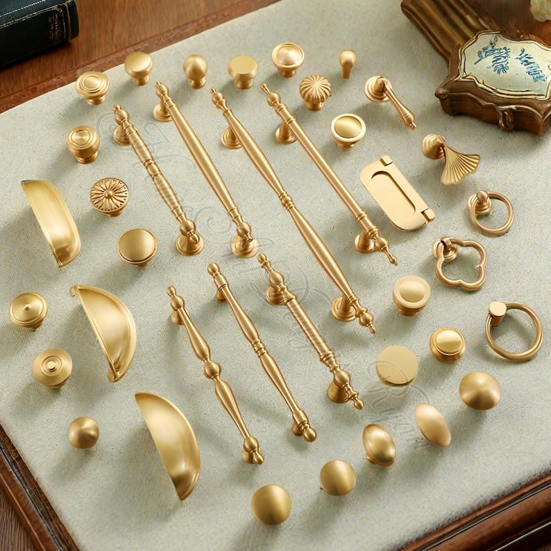 Puede incluir: Una colecci&oacute;n de herrajes dorados para armarios, que incluye pomos, tiradores y manijas, dispuestos sobre una superficie clara. Los herrajes presentan varios dise&ntilde;os, desde pomos redondos simples hasta tiradores ornamentados, ofreciendo una gama de opciones.
