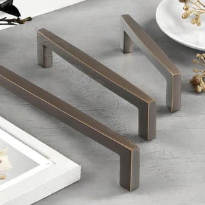 K&ouml;nnte beinhalten: Drei bronzefarbene Schrankgriffe mit einem rechteckigen Stab und quadratischen Enden. Die Hardware hat eine Vintage-&Auml;sthetik, geeignet f&uuml;r Schubladen oder Schrankt&uuml;ren. Die Griffe sind auf einer hellgrauen Oberfl&auml;che angeordnet.