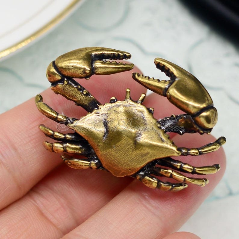 Puede incluir: Un broche de metal dorado en forma de cangrejo con garras y patas detalladas. El cangrejo est&aacute; en posici&oacute;n enroscada, con las garras levantadas.
