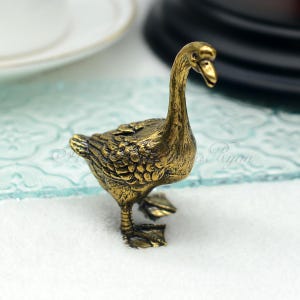 May include: A small, gold-coloured goose figurine with detailed feathers. The goose is standing on a white surface.