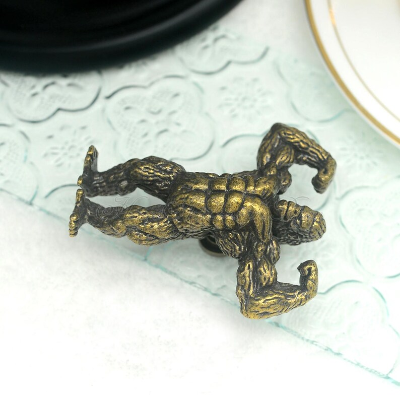 Pu&ograve; includere: Un pomello per armadio in metallo dorato a forma di gorilla muscoloso. Il gorilla &egrave; in piedi con le braccia alzate.