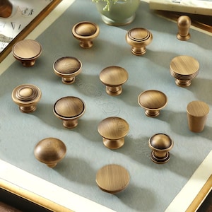 Puede incluir: Una colecci&oacute;n de tiradores de armario de lat&oacute;n antiguo en varias formas y tama&ntilde;os. Los tiradores tienen un acabado c&aacute;lido y envejecido, con dise&ntilde;os que van desde c&uacute;pulas simples hasta estilos detallados y en capas. La disposici&oacute;n sugiere una exhibici&oacute;n de opciones de hardware.