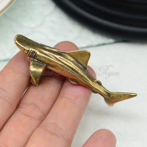 Puede incluir: Una figurita de tiburón de metal de color dorado. El tiburón está representado en una pose de natación con la boca abierta y las aletas extendidas.