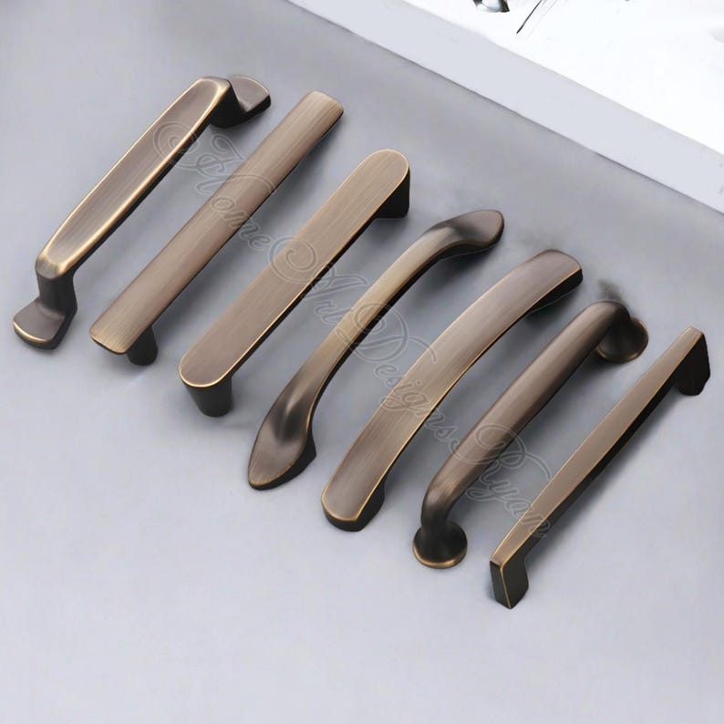 K&ouml;nnte beinhalten: Eine Sammlung von bronzefarbenen Schrankgriffen in verschiedenen Designs. Die Griffe haben eine gebogene oder rechteckige Form mit einem Vintage-Look. Die Hardware ist auf einer hellgrauen Oberfl&auml;che angeordnet, die die verschiedenen Stile zeigt.