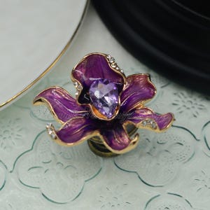 Könnte beinhalten: Ein violetter, blumenförmiger Flaschenverschluss mit einem klaren Kristall in der Mitte. Der Verschluss ist aus Metall und hat eine goldene Oberfläche.