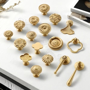 May include: A collection of various brass cabinet knobs and pulls. The knobs feature floral, geometric, and unique designs. The hardware is in a warm gold tone, with a matte finish. The assortment includes knobs, ring pulls, and handle pulls.