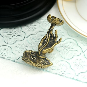 Peut inclure: Une sculpture de main en métal doré tenant une fleur de lotus. La main repose sur une base avec un motif en spirale.