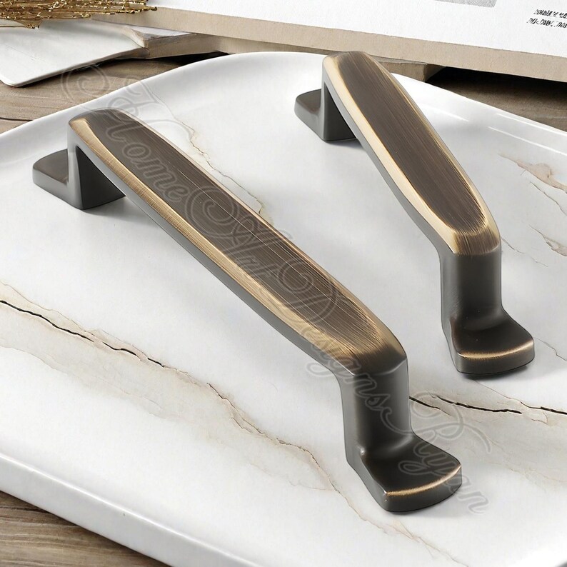 K&ouml;nnte beinhalten: Zwei bronzefarbene Schrankgriffe mit rechteckigem Design und gebogenem Griff. Die Griffe haben einen dunkelbronzenen Sockel und eine hellere bronzefarbene Mitte. Die Griffe liegen auf einer wei&szlig; marmorierten Oberfl&auml;che.