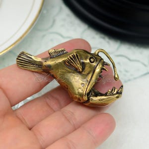 K&ouml;nnte beinhalten: Bronze-farbene Anglerfisch-Figur mit offenem Maul und scharfen Z&auml;hnen. Der Fisch hat detaillierte Flossen und einen leuchtmittelartigen K&ouml;der. Die Figur wird in einer Hand gehalten, um ihre Gr&ouml;&szlig;e und ihr detailliertes Design zu zeigen.
