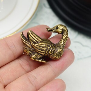 Op de afbeelding: Een klein, goudkleurig messing eendenbeeldje. De eend zit in een gekromde houding met zijn kop naar de zijkant gedraaid. Het beeldje is gedetailleerd met getextureerde veren en een klein, zwart oog.