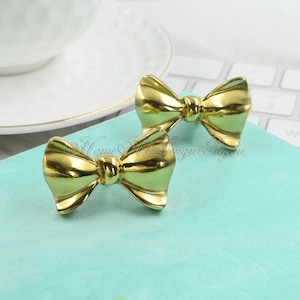May include: Two gold bow-shaped drawer pulls. The pulls are made of metal and have a shiny finish.