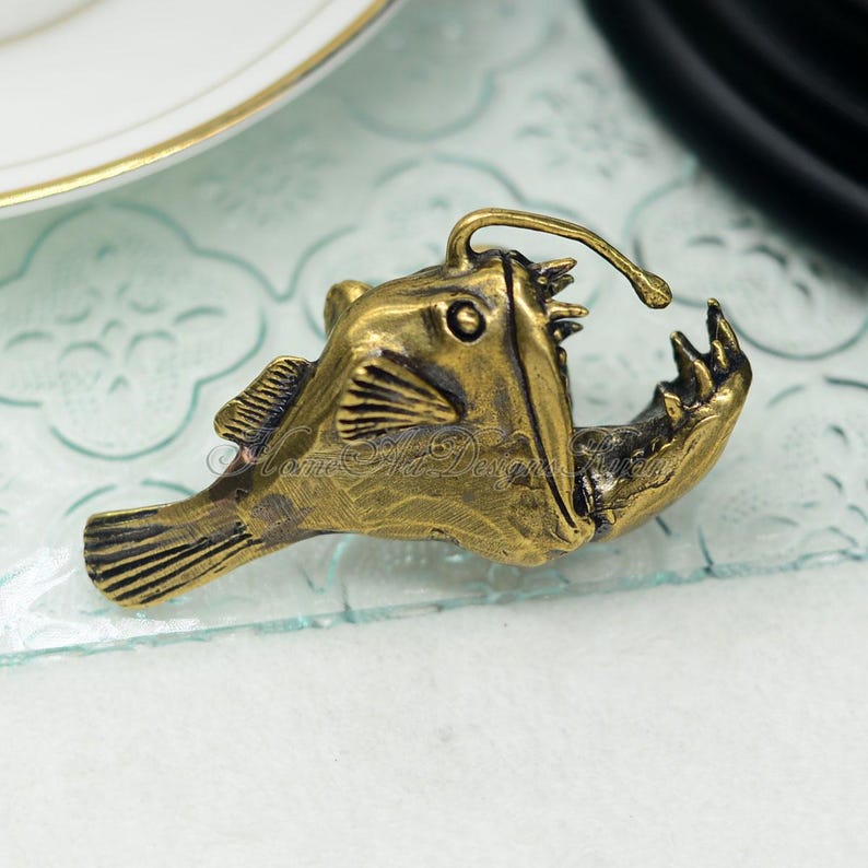 K&ouml;nnte beinhalten: Bronze-farbene Anglerfisch-Figur mit detaillierten Schuppen, Flossen und einem auff&auml;lligen K&ouml;der. Das offene Maul zeigt scharfe Z&auml;hne. Ein einzigartiges Dekorationsst&uuml;ck.