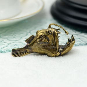 K&ouml;nnte beinhalten: Bronze farbene Anglerfisch-Figur mit offenem Maul und biolumineszierendem K&ouml;der. Die detaillierte Skulptur weist strukturierte Schuppen, Flossen und scharfe Z&auml;hne auf. Der Text "Mom's Art Designs" ist sichtbar.