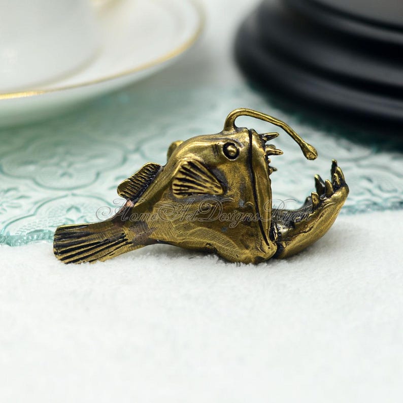 K&ouml;nnte beinhalten: Bronze farbene Anglerfisch-Figur mit offenem Maul und biolumineszierendem K&ouml;der. Die detaillierte Skulptur weist strukturierte Schuppen, Flossen und scharfe Z&auml;hne auf. Der Text "Mom's Art Designs" ist sichtbar.