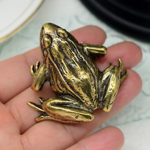 Peut inclure: Une petite figurine de grenouille en métal de couleur dorée. La grenouille est assise sur ses pattes arrière, les pattes avant tendues. La grenouille a une surface texturée et détaillée.