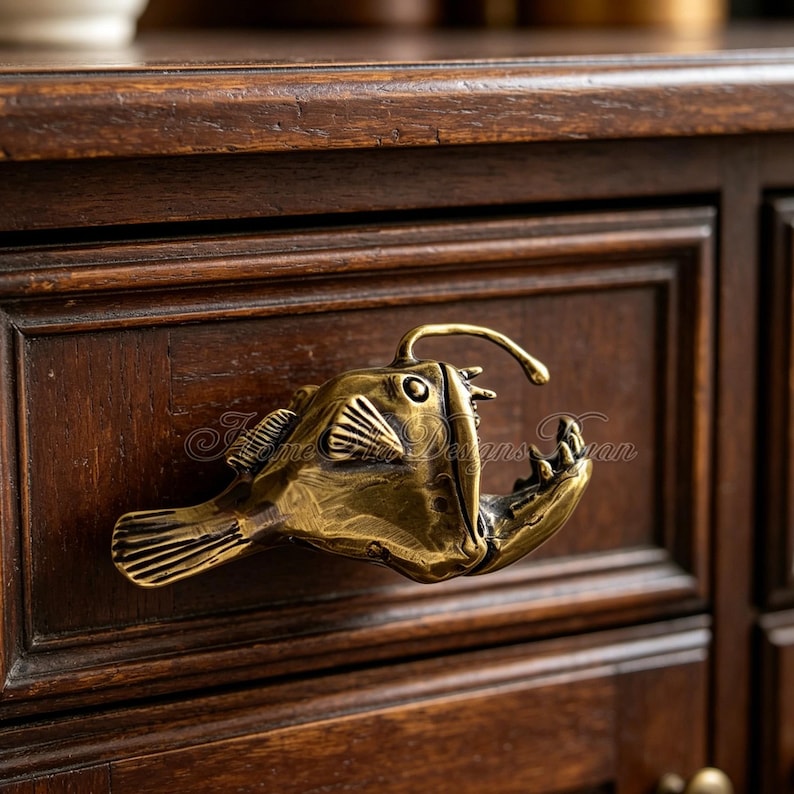 K&ouml;nnte beinhalten: Ein messingfarbener Schubladengriff in Form eines Tiefsee-Anglerfischs, montiert auf einem dunklen Holzschrank. Der Fisch hat detaillierte Flossen, ein offenes Maul und einen gebogenen K&ouml;der. Der Schrank hat ein eingelassenes Paneeldesign.