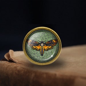 May include: A round, brass-colored pin with a glass dome featuring a detailed illustration of a Death's-head Hawkmoth with orange and black wings against a green background.