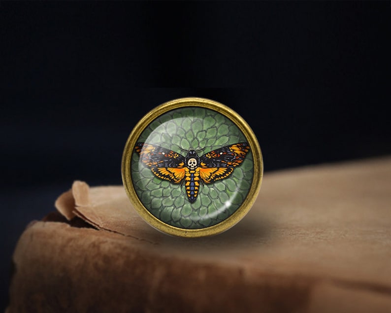 May include: A round, brass-colored pin with a glass dome featuring a detailed illustration of a Death's-head Hawkmoth with orange and black wings against a green background.