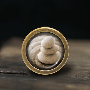 May include: A brass-colored round pendant with a glass insert featuring a stack of three smooth, light gray stones on a sandy surface.