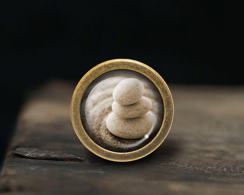 Pode incluir: Um pingente redondo de cor lat&atilde;o com um inserto de vidro que apresenta uma pilha de tr&ecirc;s pedras lisas de cor cinza claro sobre uma superf&iacute;cie arenosa.