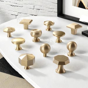 Puede incluir: Una colecci&oacute;n de tiradores dorados para armarios en varias formas geom&eacute;tricas, incluyendo esferas, oct&aacute;gonos y dise&ntilde;os de tapa plana. Los tiradores est&aacute;n dispuestos sobre una superficie blanca, mostrando sus diferentes estilos y acabados. Estas piezas de ferreter&iacute;a decorativas son adecuadas para muebles y decoraci&oacute;n del hogar.