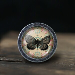 May include: A round, grey metal knob with a glass insert featuring a black butterfly on a floral pattern background.